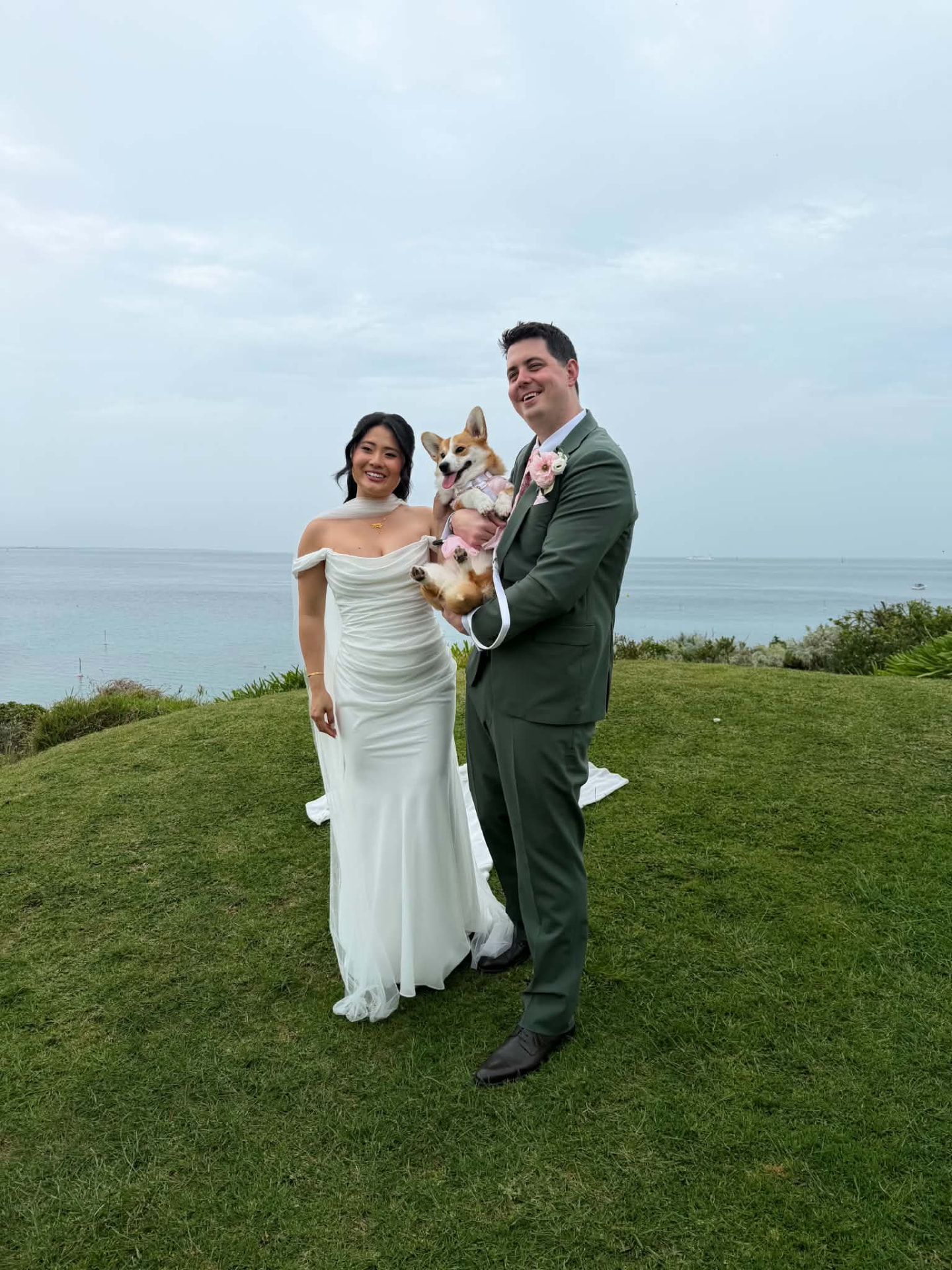 Bride and groom holding their Corgi in a pink harness at their seaside wedding