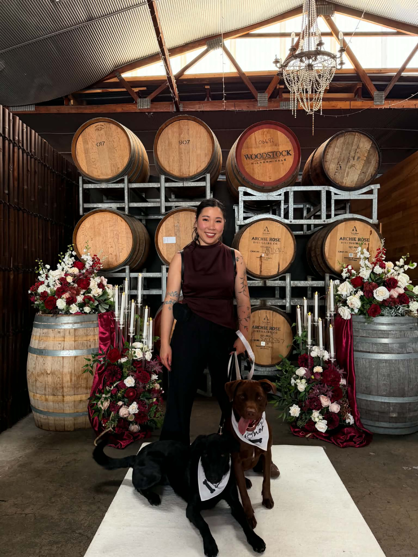 Alexandra with two dogs wearing wedding bandanas beside ceremony flowers and wine barrels